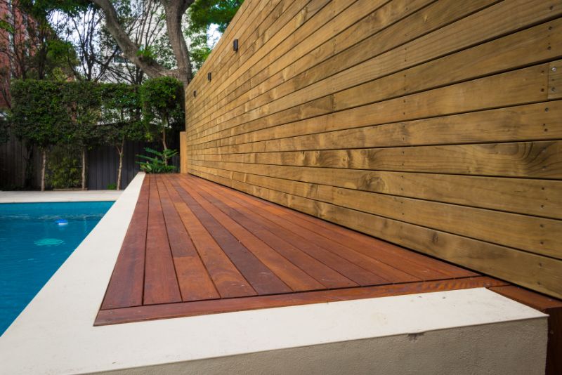 Timber deck platform beside a swimming pool, framed by a horizontal timber slat wall