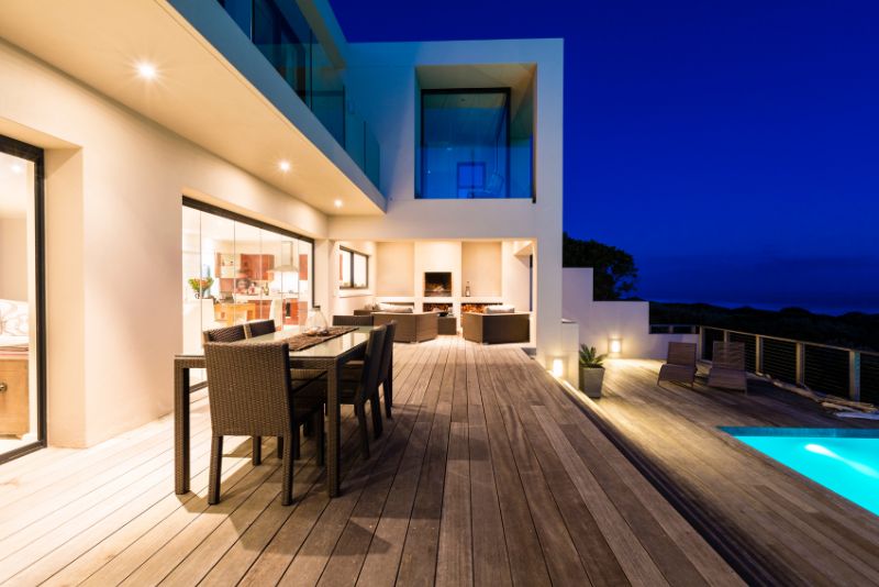 Timber pool deck with an outdoor dining table, lit at night beside an infinity pool